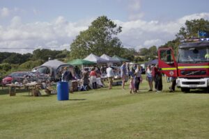 Photo of a Fire Engine in front of busy stalls at a fete, on a village green with blue cloudy sky